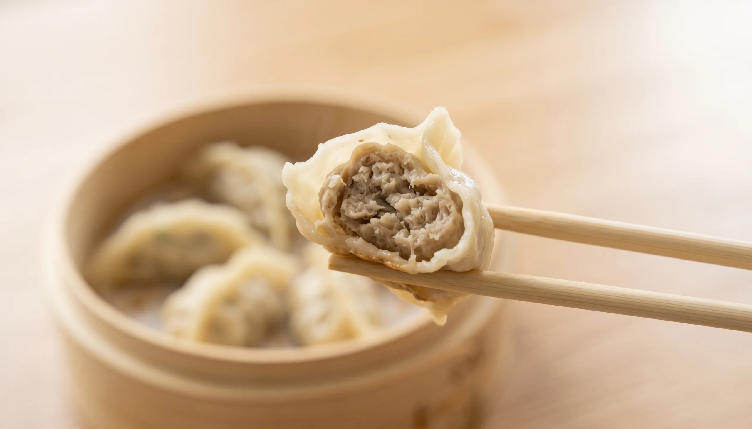 Gyoza japonesa al vapor con base dorada sostenida por palillos de madera, sobre una mesa con cuencos de salsa desenfocados